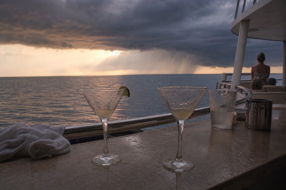Cocktail glasses on a boat bar counter with evening light on the water behind, no text, no logos, no people