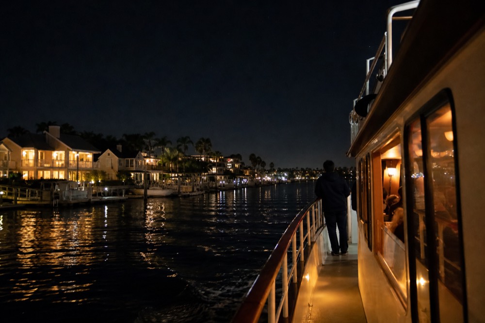 A small dinner cruise boat passing lit-up waterfront homes at night, no text, no logos, no people