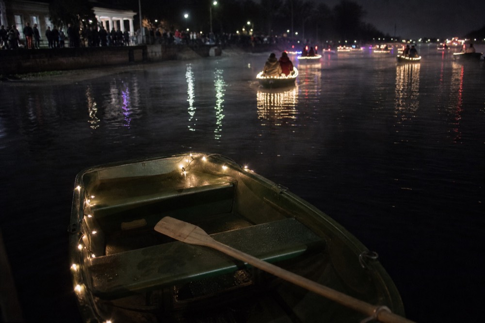 Rowboats with string lights drifting on a dark river before the main parade, no text, no logos, no people.
